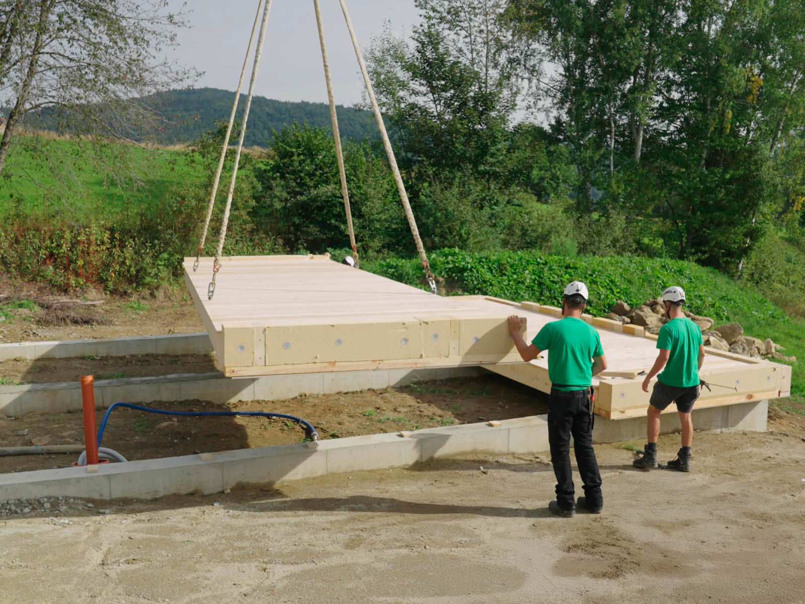 Eine Holzbodenplatte wird per Kran mit Seilen auf einer Baustelle auf Streifenfundamente gelegt. Zwei Mitarbeiter von Brunthaler arbeiten an der richtigen Platzierung der Holzbodenplatte. © Brunthaler Naturhausmanufaktur