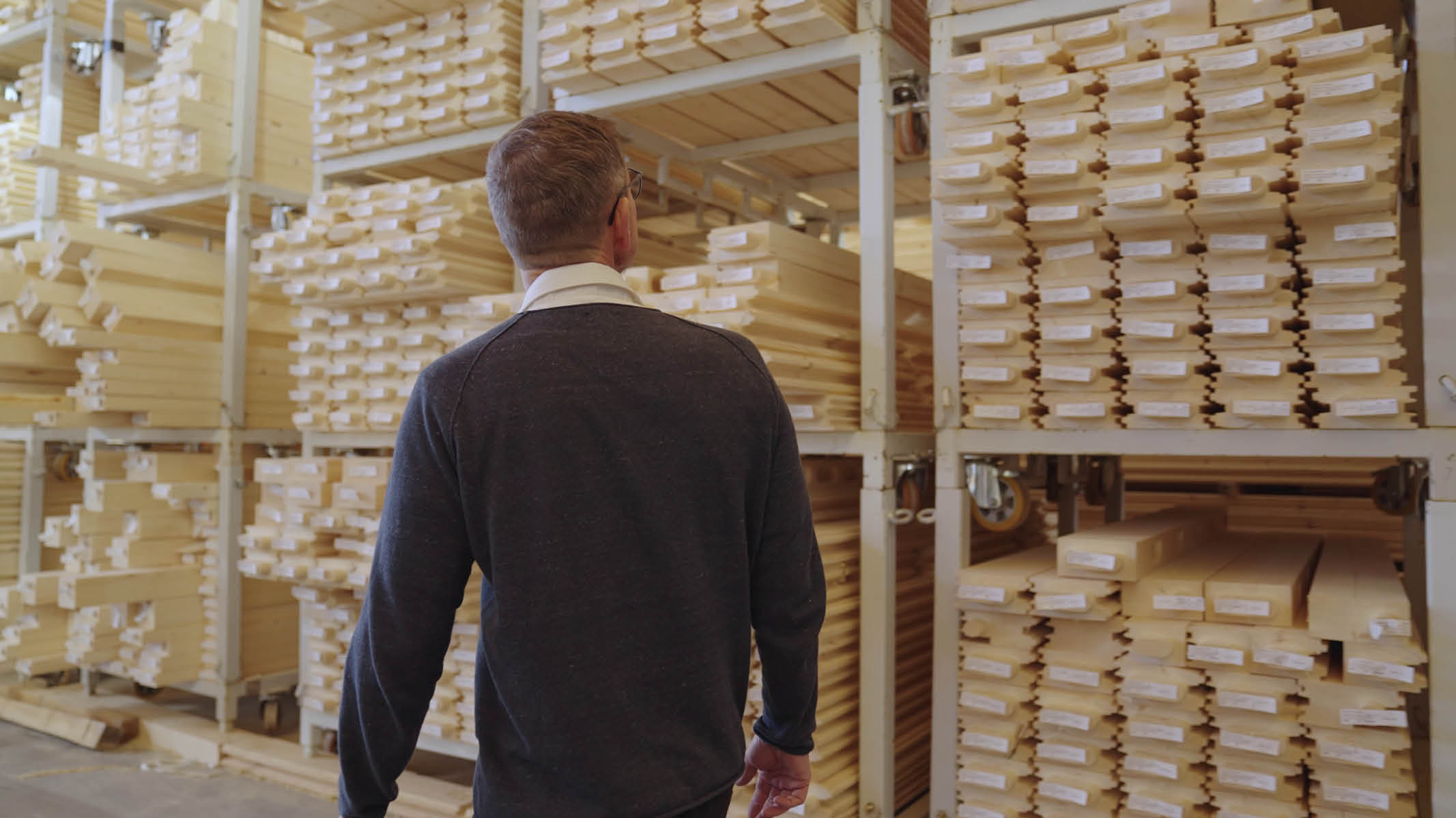 Ein Mitarbeiter geht durch eine Halle auf dem Werksgelände von Brunthaler und blickt auf Reihen von großen Lagerregalen mit vielen MONOBLOCK-Balken, die jeweils mit einem kleinen Stück Papier beschriftet sind. © Brunthaler Naturhausmanufaktur