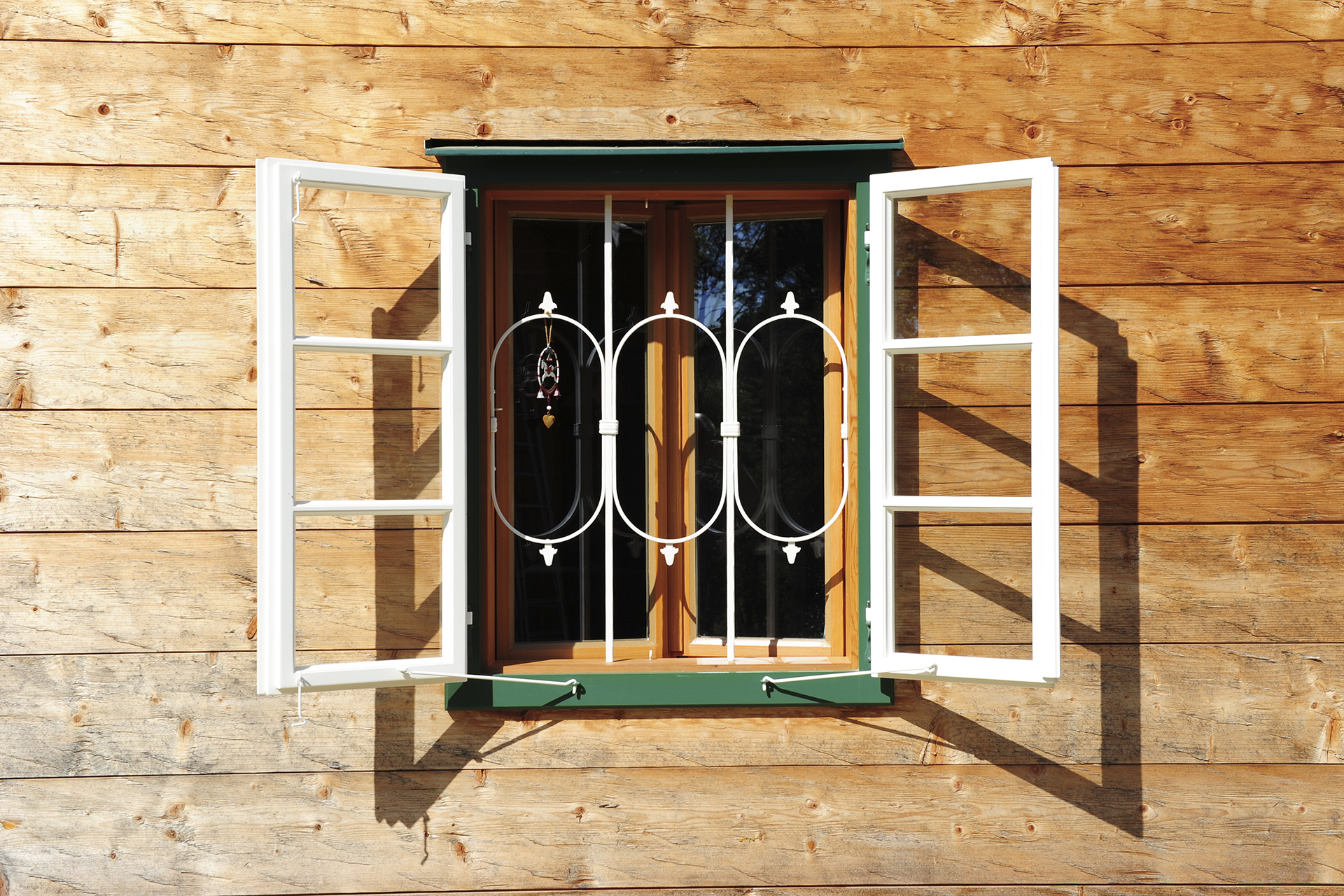 Detailaufnahme einer Blockbohlenfassade mit grün gerahmten Fenstern mit weißen dekorativen und sehr filigranen Fenstergittern und weißen Fensterläden. © Brunthaler Naturhausmanufaktur