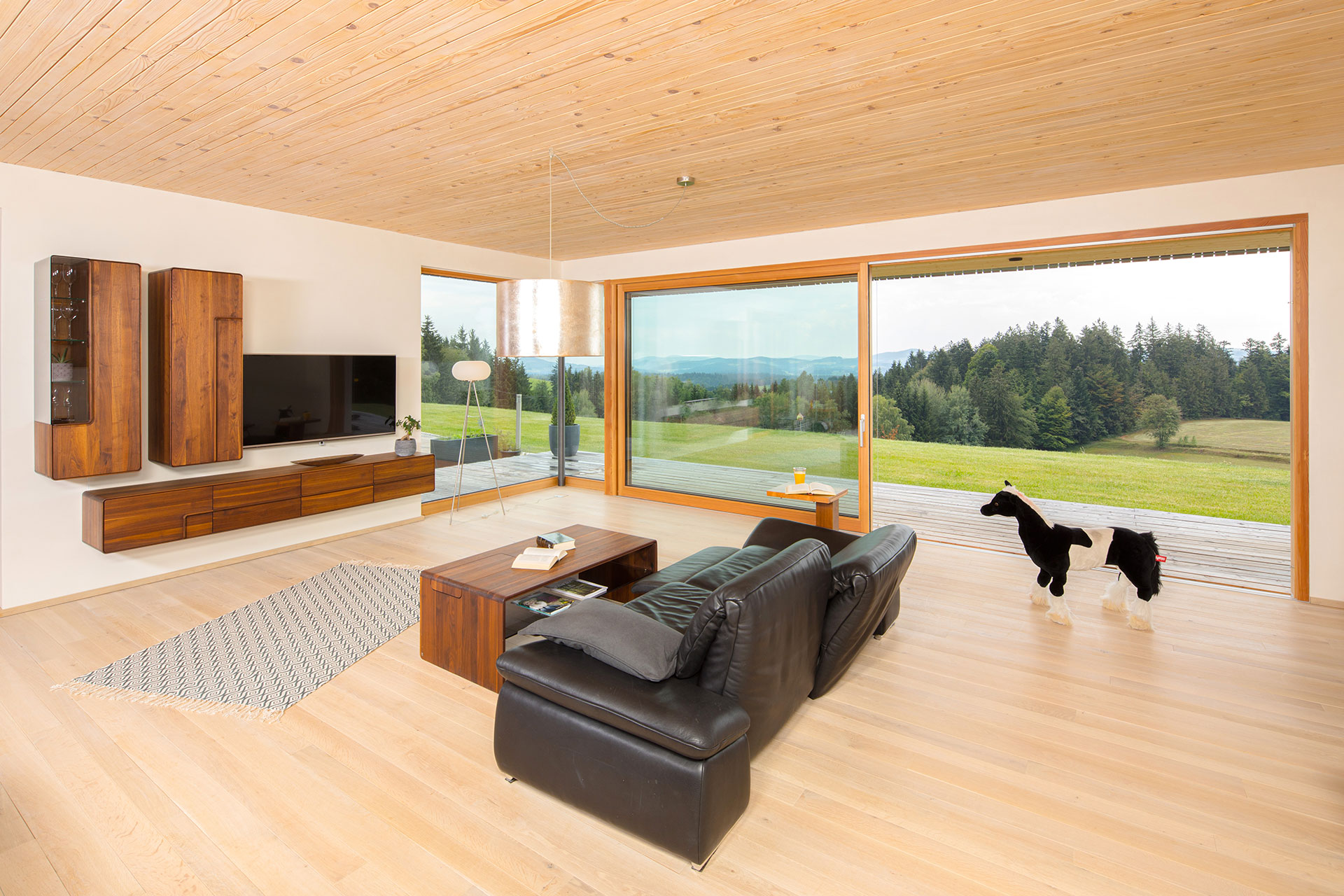 Großzügiges Wohnzimmer mit großer Glasfront und Terrassentür, Holzdecke, Holzfußboden und verputzten Wänden. Schwebende Fernsehwand aus dunklem Holz, schwarze Ledercouch und ein Spielzeugpferd. © Brunthaler Naturhausmanufaktur