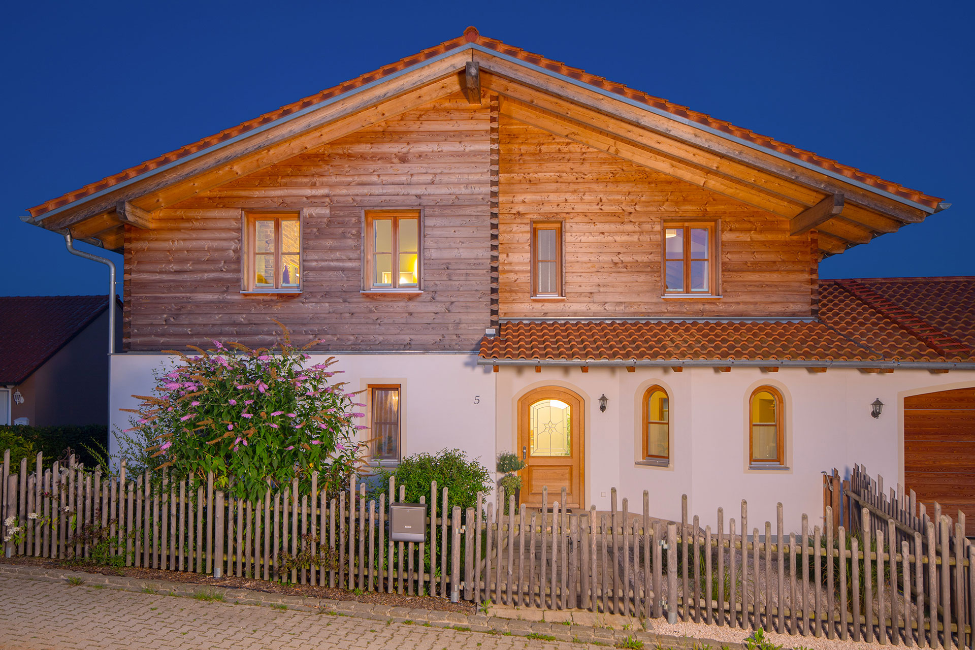 Abendstimmung eines großzügigen Massivholz-Landhauses mit Blockbohlenfassade oben und verputzter Fassade im Erdgeschoss. Rustikaler Zaun umsäumt das Grundstück. © Brunthaler Naturhausmanufaktur
