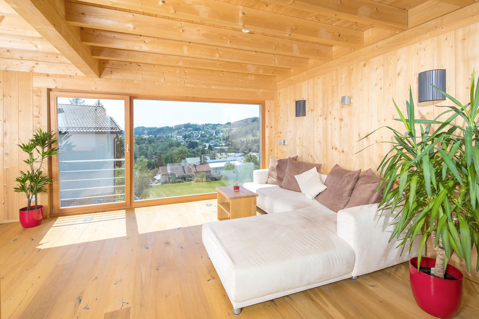 Wohnzimmer in einem Massivholzhaus mit weißem Sofa, Sichtholzdecke und bodentiefen Fenstern. © Brunthaler Naturhausmanufaktur