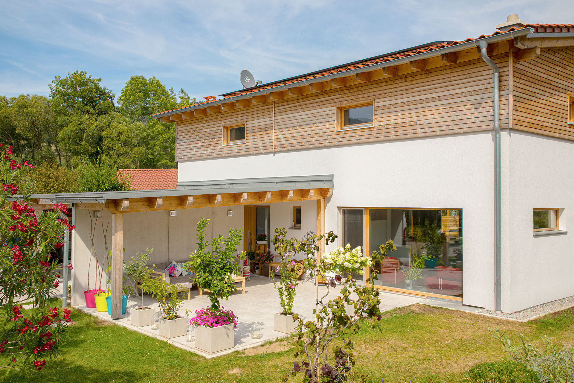 Klassisches Einfamilienhaus mit verputzter Fassade und einem kleinen Teil an horizontaler Holzfassade. Große Fenster und eine betonierte Terrasse mit Überdachung ergänzen das Holzhaus. © Brunthaler Naturhausmanufaktur