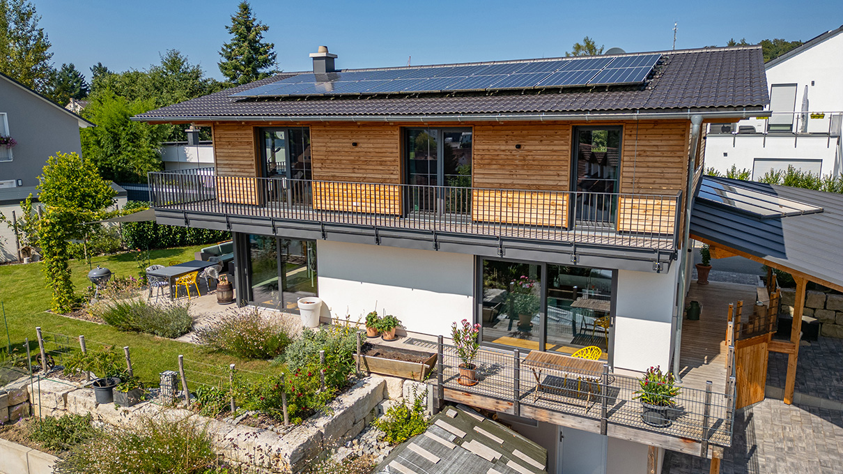 Klassisches Einfamilien-Holzhaus mit umlaufendem Balkon und dunkelgrauen Dachschindeln in Hanglage. Oben Holzfassade und unten verputzt. © Brunthaler Naturhausmanufaktur