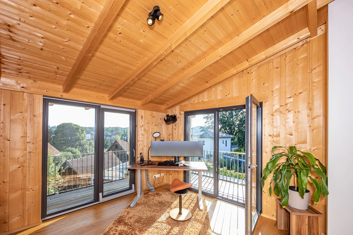 Home Office Büro mit Schreibtisch und großem Monitor. Der Raum im Holzhaus hat unbehandelte Holzwände und eine Holzdecke. Der Schreibtisch steht im Eck zwischen zwei Balkontüren, wodurch der Raum hell und lichtdurchflutet ist. © Brunthaler Naturhausmanufaktur