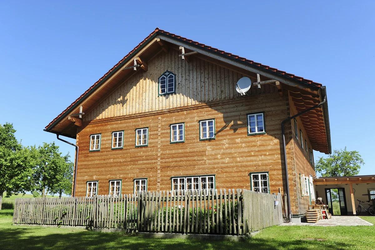 Innviertler Hof aus Massivholz mit traditioneller Blockbohlenfassade, weiß verputztem Erdgeschoss und rotem Satteldach inmitten einer grünen Wiesenlandschaft. © Brunthaler Naturhausmanufaktur