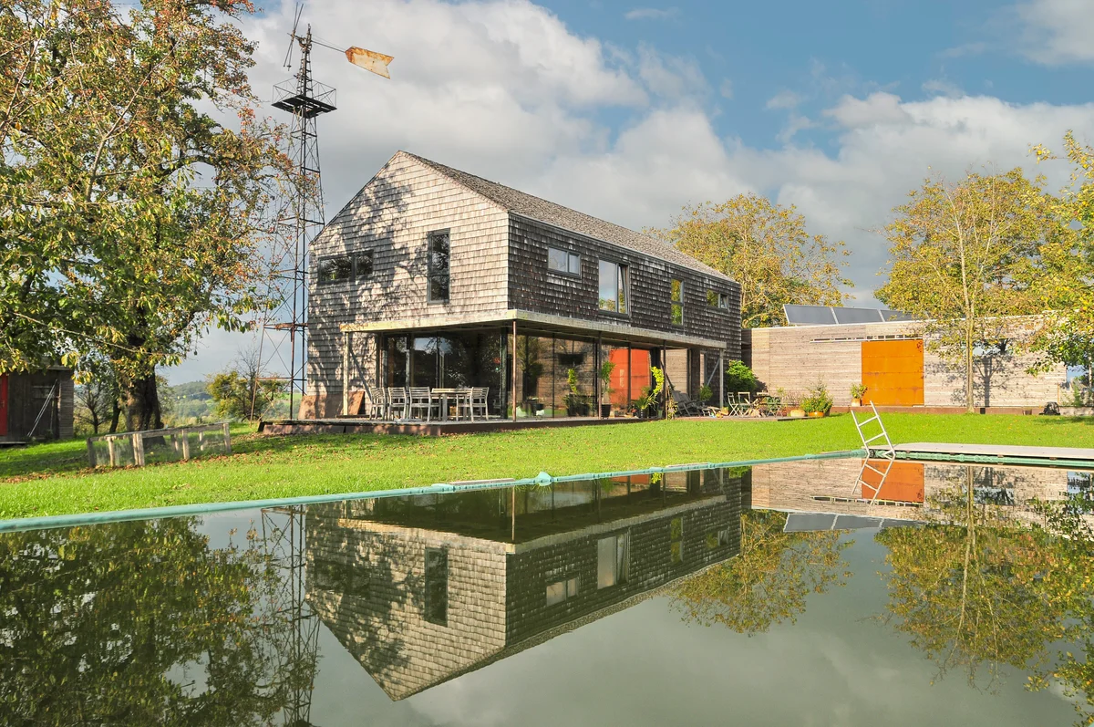 Modernes Massivholzhaus mit vergrauter Holzschindelfassade ohne Dachüberhang. Davor sieht man einen grünen Rasen mit abgedecktem Pool und dahinter steht eine sehr große Windmühle aus Metall. © Brunthaler Naturhausmanufaktur