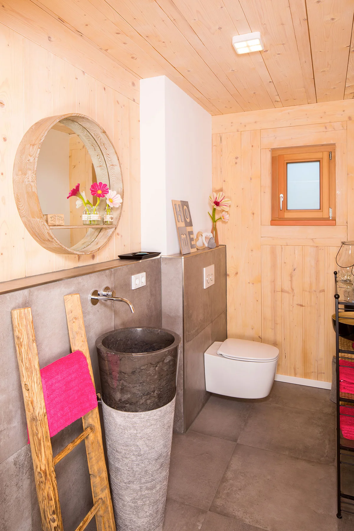 Modernes und kreatives Klo mit Holzwänden und grauen Fliesen und einem rustikalen runden Spiegel mit pinken Farbakzenten durch Blumen und Handtücher. Das Waschbecken ist trichterförmig und freistehend aus einem dunklen und hellen Steinmaterial. © Brunthaler Naturhausmanufaktur