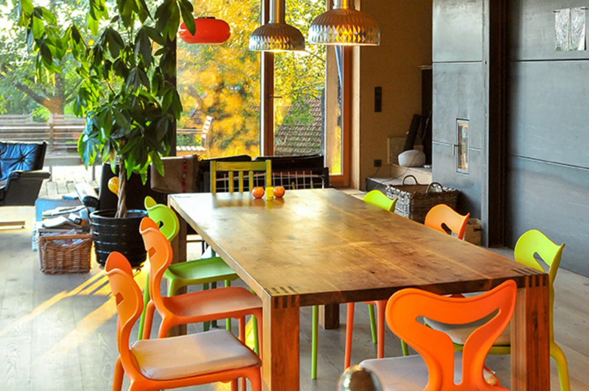 Offener Ess- und Wohnbereich mit verschiedenen, bunten Stühlen und dunklem, großflächigem Zentralofen, der in die Wand des Massivholzhauses integriert ist. © Brunthaler Naturhausmanufaktur