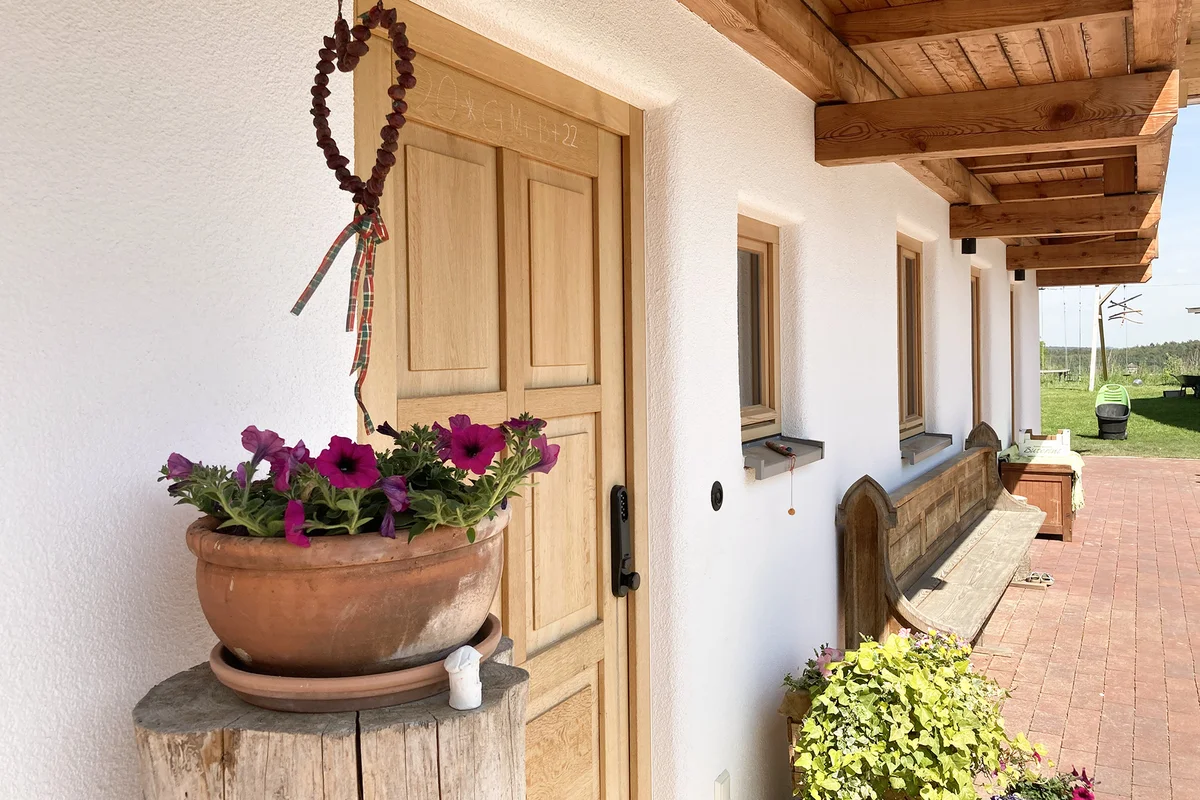 Detailansicht der Eingangstür aus Holz. Die verputzte Fassade hat abgerundete Ecken. Ein Blumentopf steht auf einem Baumstamm vor dem Massivholzhaus. © Brunthaler Naturhausmanufaktur