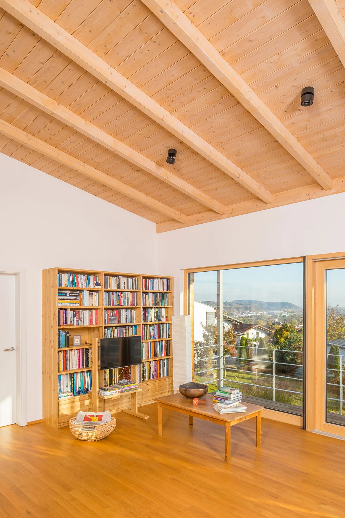 Lese- und Fernsehbereich im Holzhaus mit hoher Decke und Sichtdachstuhl. Der helle Raum hat weiße Wände und eine Balkontür mit Blick auf die Umgebung. Ein Holzregal gefüllt mit Büchern, ein Fernseher mit Holzfüßen und ein kleiner niedriger Tisch machen den Eckbereich zur kleinen Bibliothek. © Brunthaler Naturhausmanufaktur