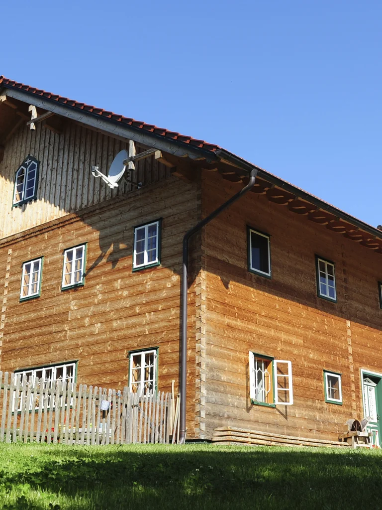 Innviertler Hof aus Massivholz mit traditioneller Blockbohlenfassade, weiß verputztem Erdgeschoss und rotem Satteldach inmitten einer grünen Wiesenlandschaft. © Brunthaler Naturhausmanufaktur