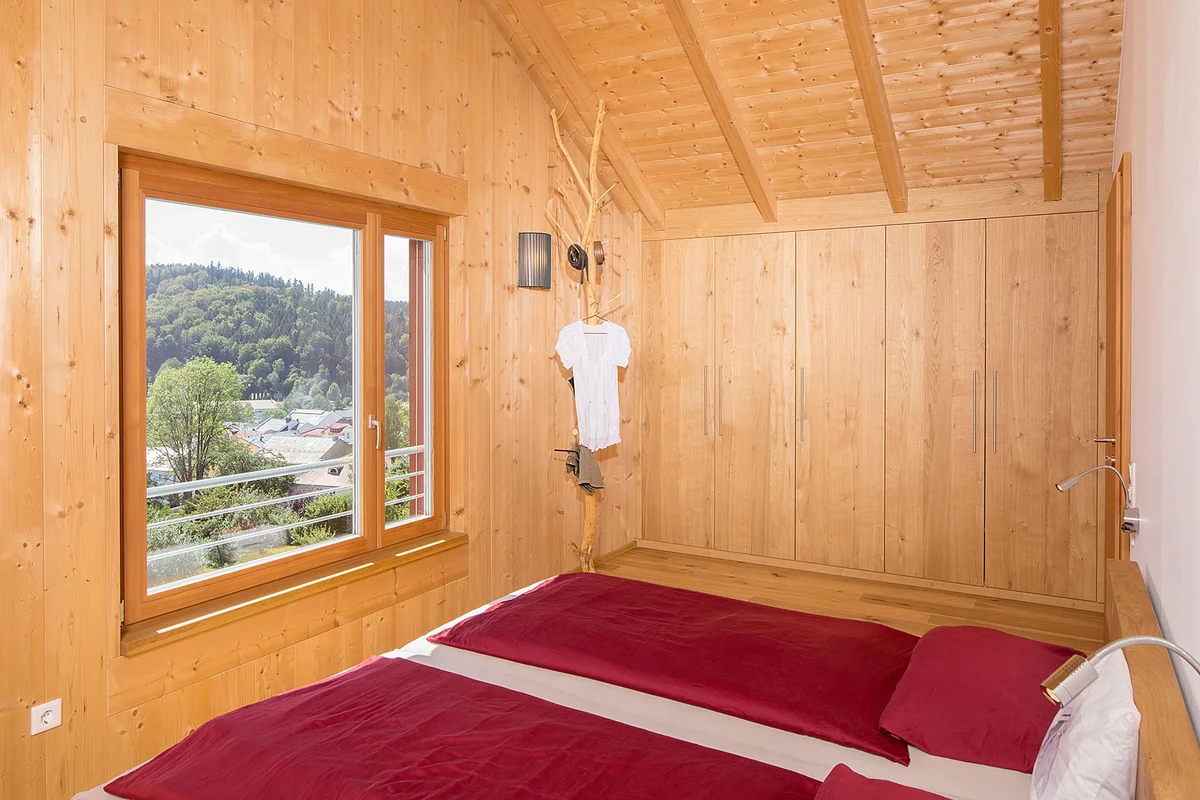 Schlafzimmer in einem Massivholzhaus mit Sichtholzwänden und Sichtholzdecke, einem großen Fenster. Im Eck steht ein Ast, der an Boden und Decke angemacht als rustikaler und kreativer Kleiderständer fungiert. © Brunthaler Naturhausmanufaktur