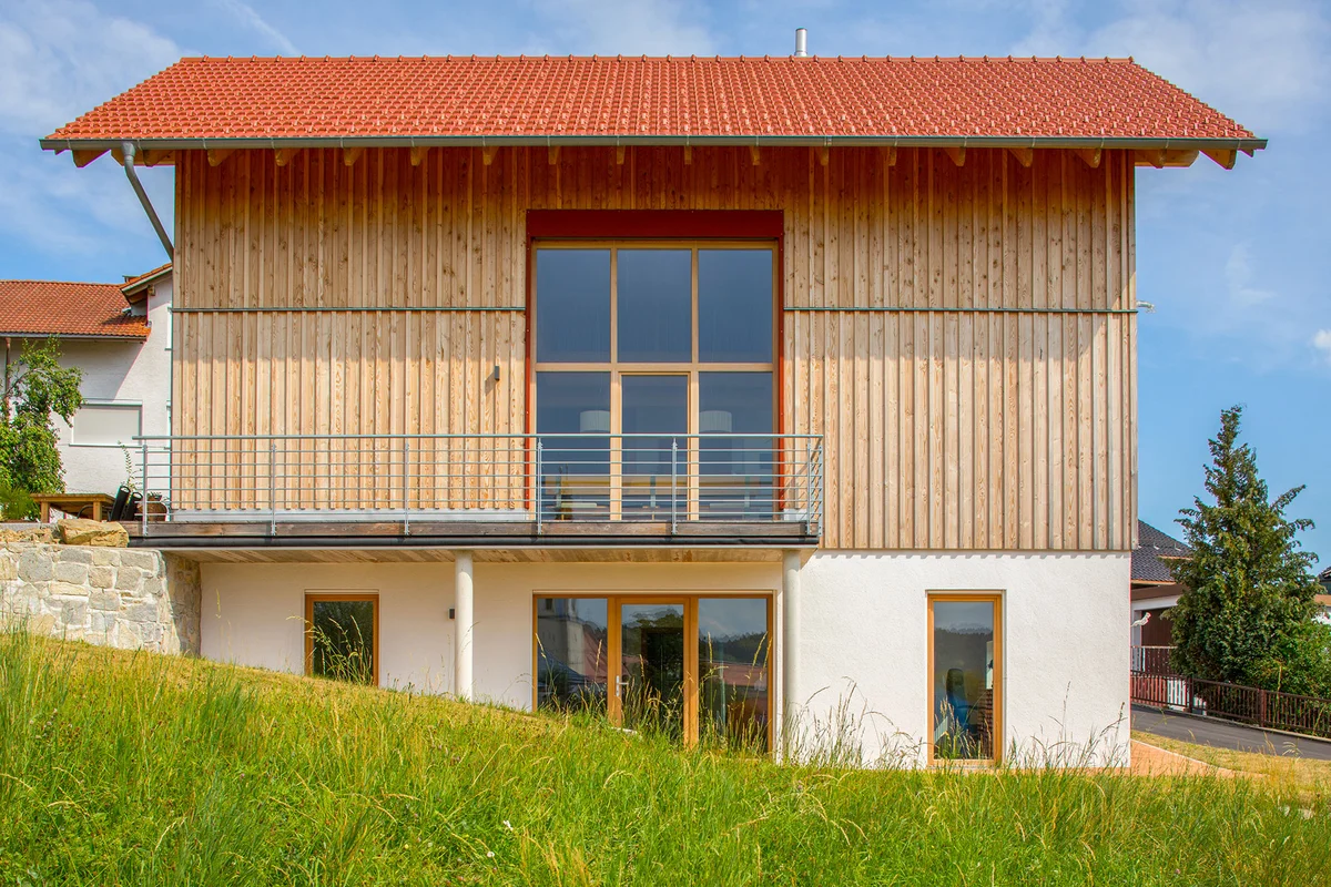 Modernes Holzhaus in Hanglage mit vertikaler Holzfassade, roten Farbakzenten, einer Holzterrasse, die sich seitlich um das Haus zieht. Der untere Bereich der Fassade ist weiß verputzt. Seitlich hat das Haus ein mehrstöckiges Fenster. Das Haus hat ein rotes Satteldach. © Brunthaler Naturhausmanufaktur