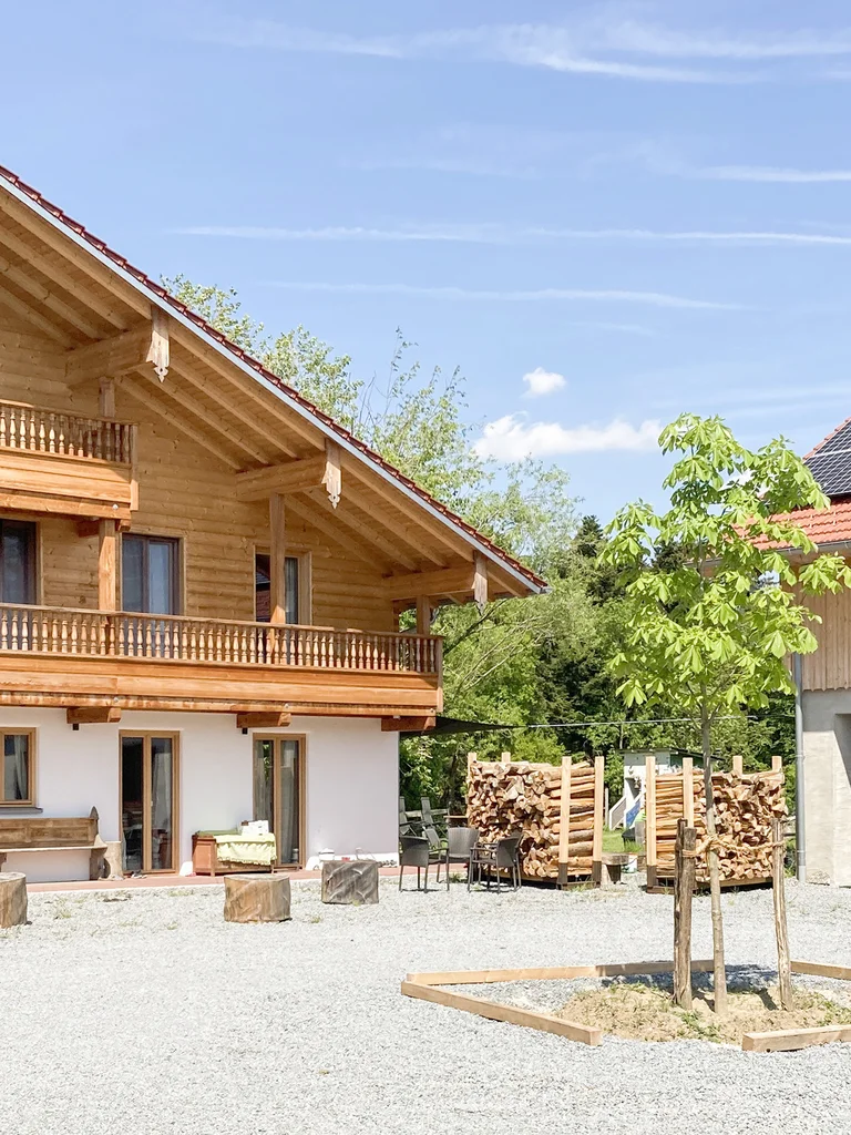 Großes bayerisches Landhaus. Teilweise weiß verputzt und oben mit rustikaler Blockbohlenfassade und zwei großzügigen Balkonen. Rechts daneben steht ein moderneres Haus mit Photovoltaik-Panels auf dem Dach. © Brunthaler Naturhausmanufaktur