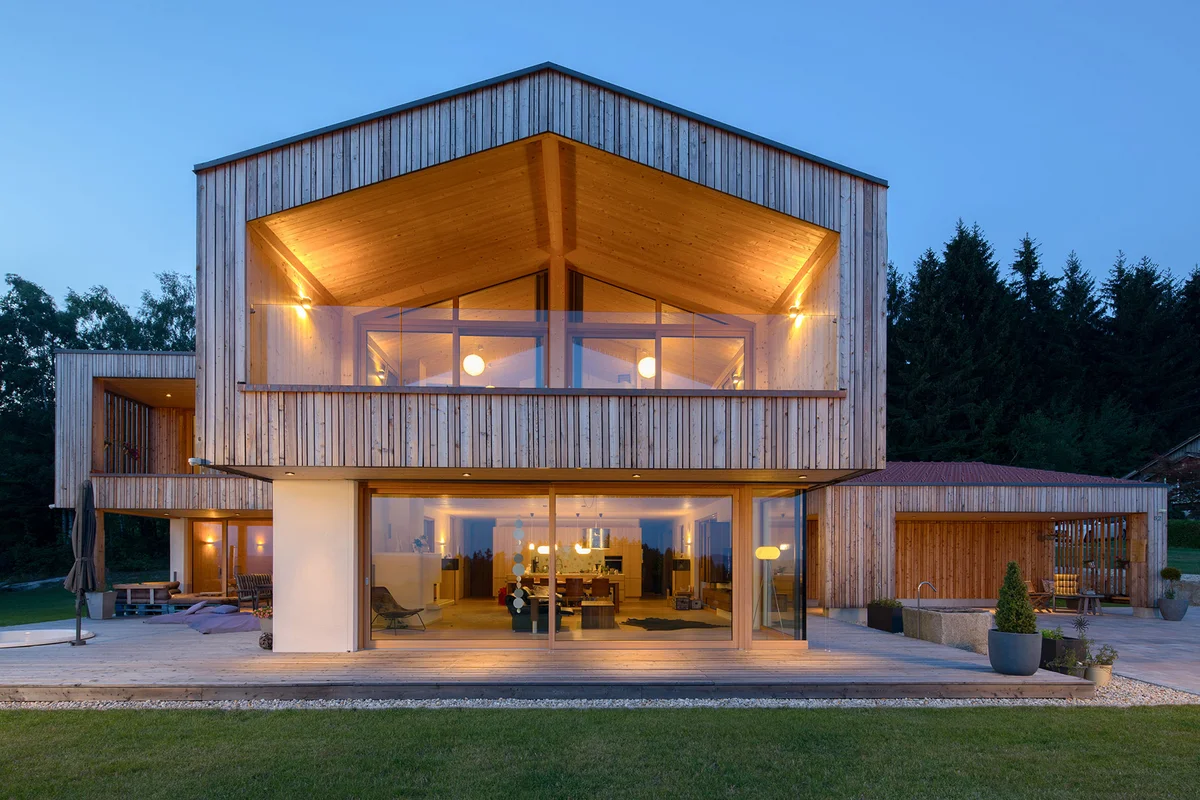Abendstimmung eines modernen Architektenholzhauses mit vertikaler heller Holzfassade und verputztem Erdgeschoss. Ein großer überhängender Balkon mit Glasgeländer liegt über einer großen Glasfront im Erdgeschoss. © Brunthaler Naturhausmanufaktur