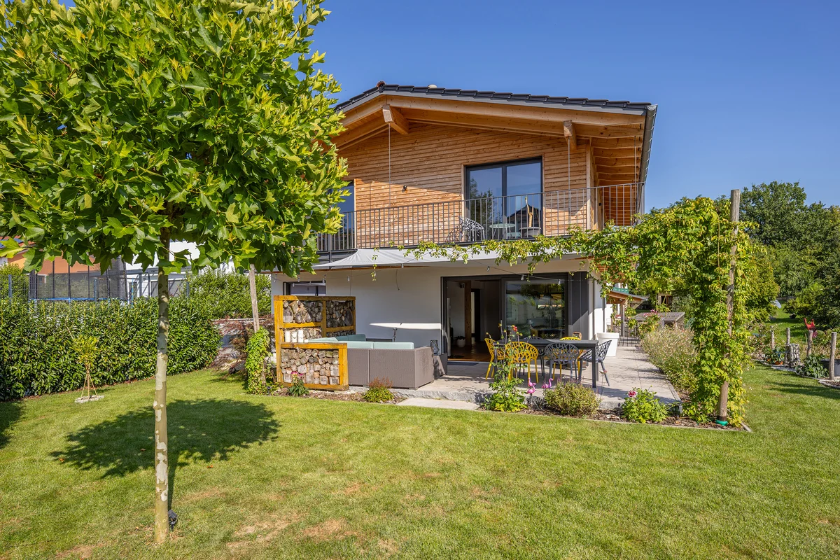 Klassische Einfamilien-Holzhaus mit umlaufendem Balkon in Hanglage. Oben Holzfassade unten verputzt. Gemütliche Terrasse mit Raumteiler aus gestapeltem Stückholz und Kletterwein, der sich zum Balkon hochrankt. Im Vordergrund ist der Garten mit einem kleinen Baum. © Brunthaler Naturhausmanufaktur