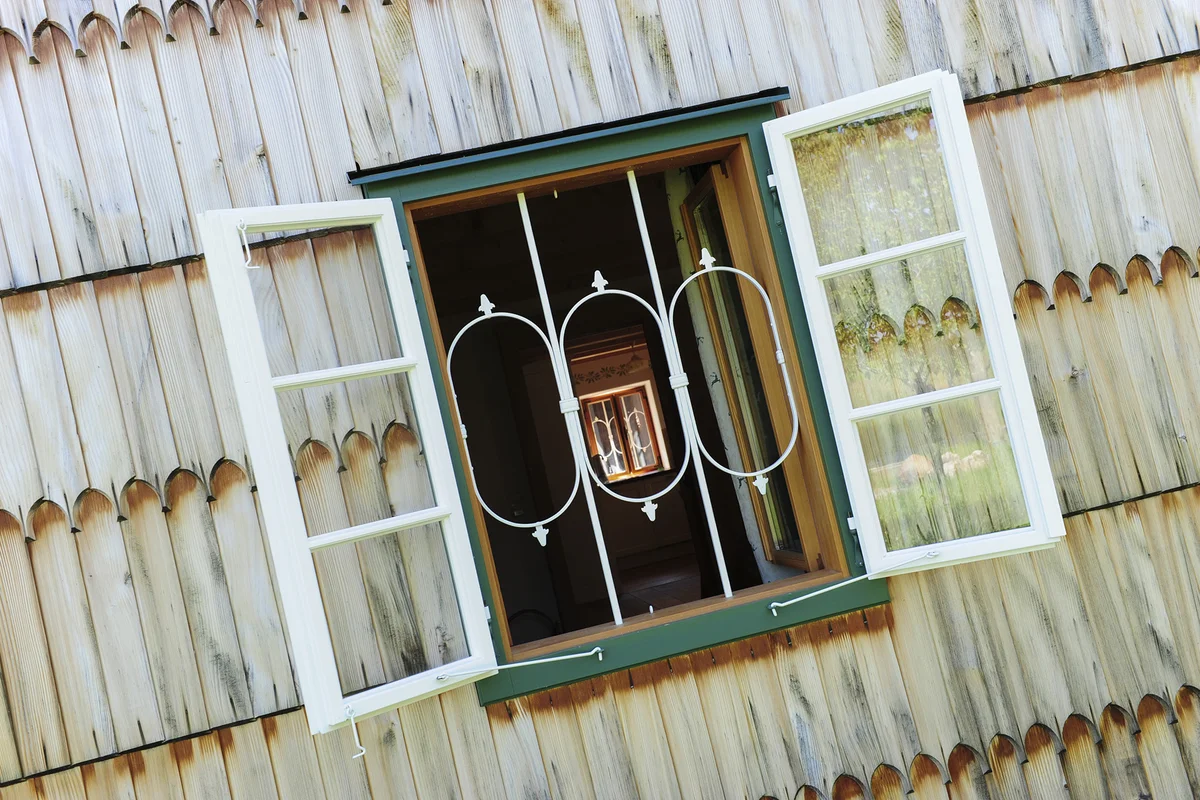 Detailaufnahme einer Holzschindelfassade mit grün gerahmten Fenstern mit weißen dekorativen und sehr filigranen Fenstergittern und weißen Fensterläden. © Brunthaler Naturhausmanufaktur