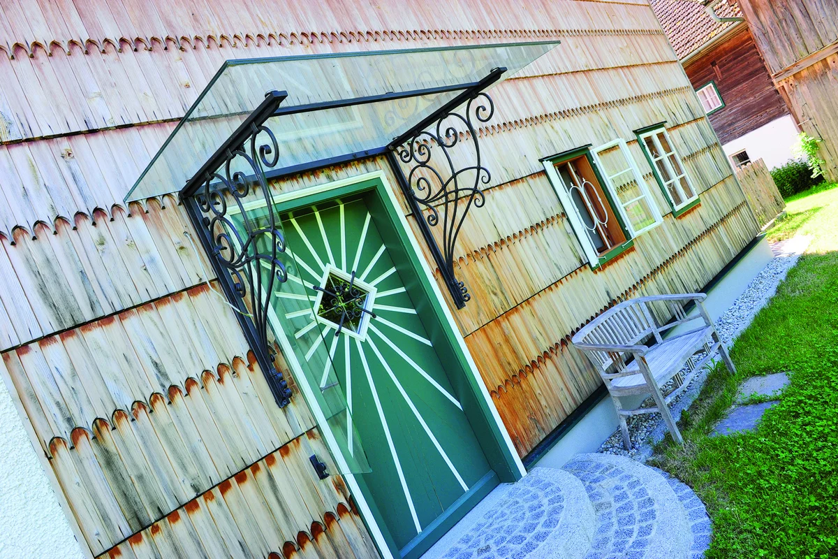 Grüne Haustür mit kleinem weiß umrandeten Fenster, das von weißen Streben umrandet ist, als wären es Strahlen. Ein kleines Glasdach mit filigranen, verschnörkelten Eisenhalterungen ist über der Tür an einer urigen Holzschindelfassade angemacht. © Brunthaler Naturhausmanufaktur