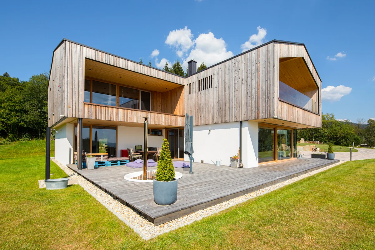 Modernes Architektenholzhaus mit überhängendem Stockwerk, ohne Dachüberstand, mit geradlinigen großen Balkonen und einer üppigen Holzterrasse. Vertikale Holzfassade und verputzte Fasse im Erdgeschoss. © Brunthaler Naturhausmanufaktur