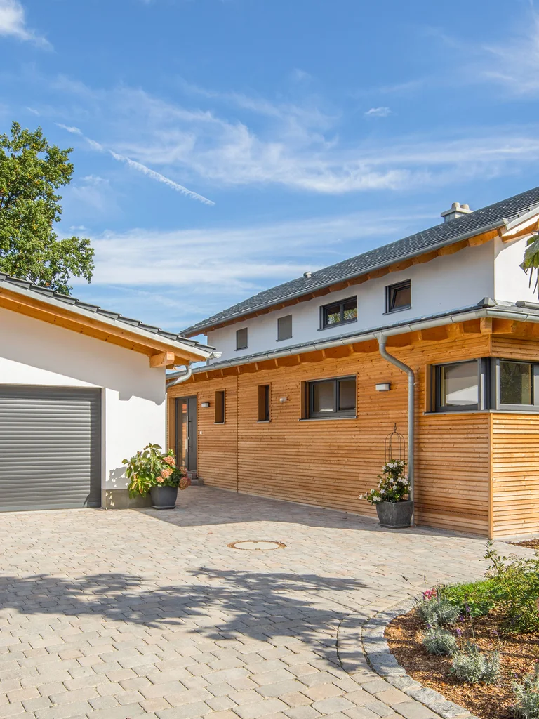 Einfamilienhaus mit Garage. Das Holzhaus ist teilweise verputzt und hat einen überdachten Übereck-Erker mit einer horizontalen Holzfassade. Auf der anderen Seite ist eine überdachte Terrasse. © Brunthaler Naturhausmanufaktur