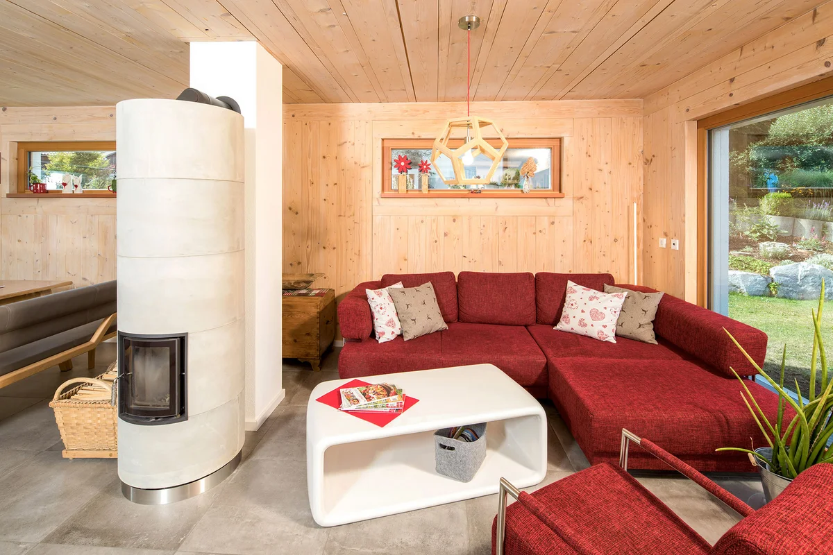 Wohnbereich mit dunkelroter Couch und einem Holzofen in Betonoptik stehen vor einer Holzwand im Einfamilienhaus. © Brunthaler Naturhausmanufaktur