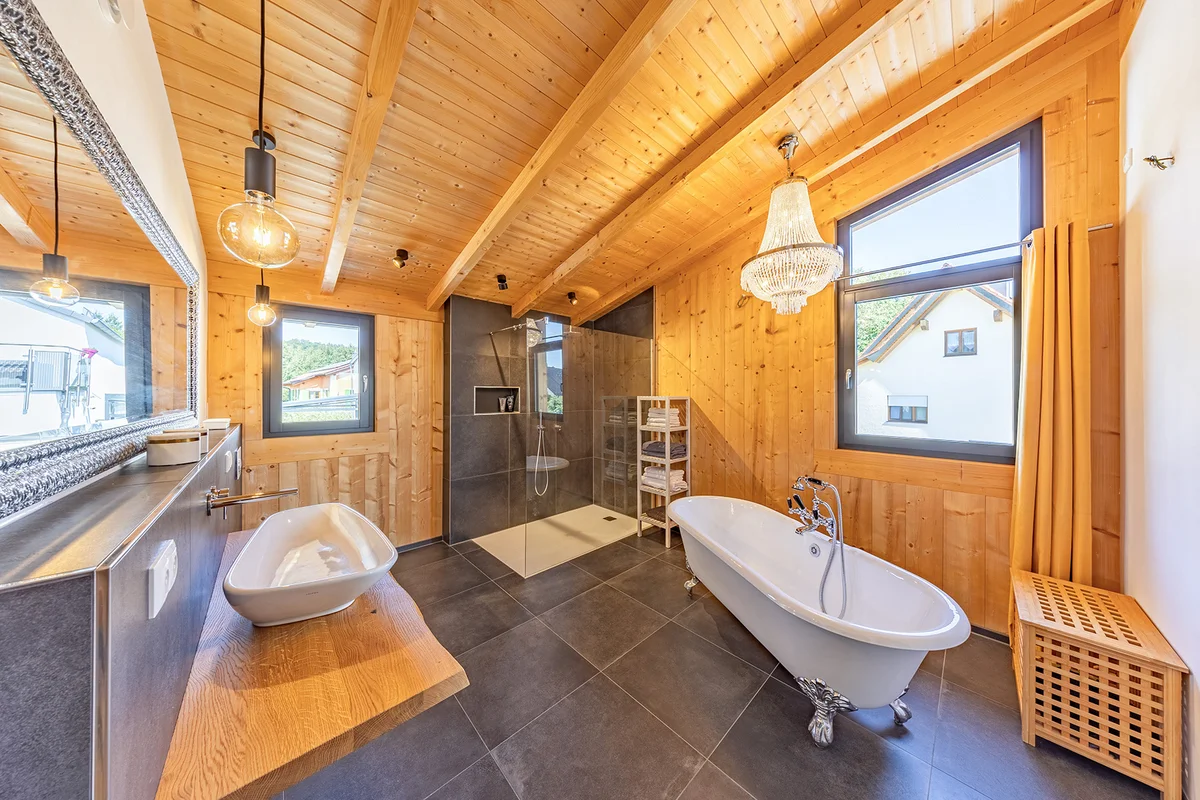 Geräumiges Badezimmer im Holzhaus mit dunkelgrauen großen Fließen und Holzwänden. Eine Badewanne mit silbernen Füßen steht freistehend im Raum. Ein funkelnder Kronleuchter hängt über der Badewanne. Der Raum wirkt warm. © Brunthaler Naturhausmanufaktur