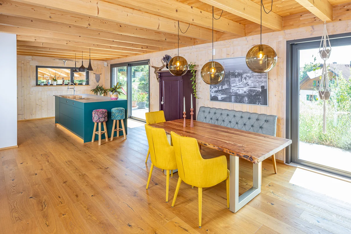Küche und Essbereich im Holzhaus. Die Wände sind eine MONOBLOCK-Wand in hellem Fichtenholz. Eine auffällige große zentrale Kücheninsel in Petrolblau, ein Schrank in dunklem Lila und Stühle in Sonnenblumengelb lassen den Trend "color blocking" in das Holzhaus einziehen. © Brunthaler Naturhausmanufaktur