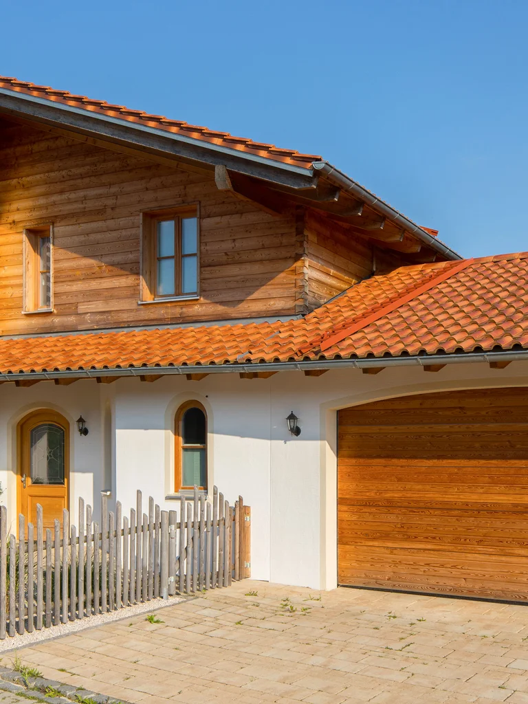 Großzügiges Massivholz-Landhaus mit Blockbohlenfassade oben und verputzter Fassade im Erdgeschoss. Rustikaler Zaun umsäumt das Grundstück. © Brunthaler Naturhausmanufaktur