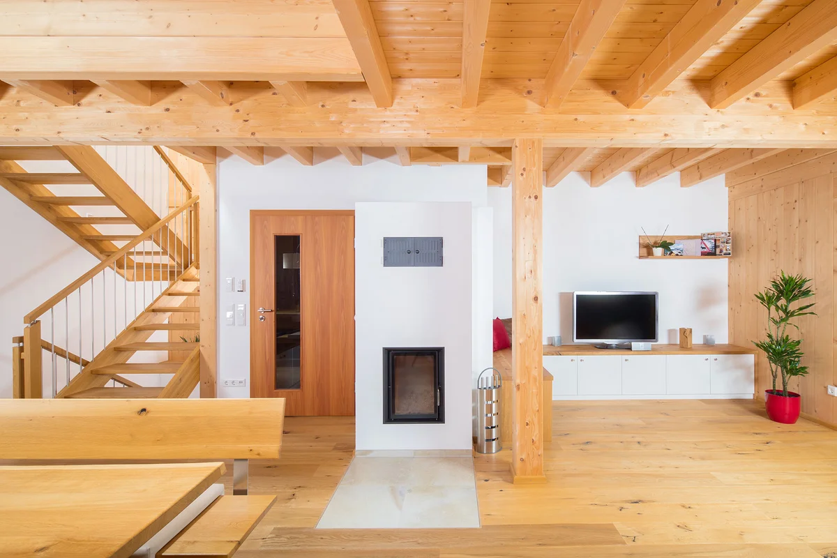 Wohn- und Essbereich mit großen zentralen Holzofen, einer Treppe in die obere Galerie. Die Holzbalkendecke und Stützbalken sind gut sichtbar. © Brunthaler Naturhausmanufaktur