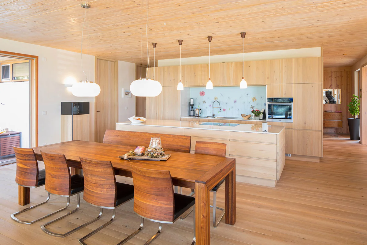 Moderne Küche mit flachen hellen Holzfronten, einem Esstisch mit Stühlen aus warmem rot-orangem Holz. © Brunthaler Naturhausmanufaktur
