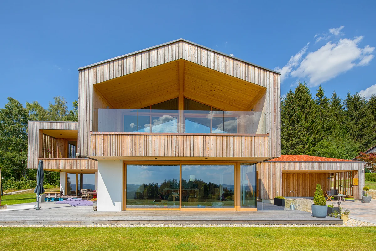 Modernes Architektenholzhaus mit vertikaler heller Holzfassade und verputztem Erdgeschoss. Ein großer überhängender Balkon mit Glasgeländer liegt über einer großen Glasfront im Erdgeschoss. © Brunthaler Naturhausmanufaktur