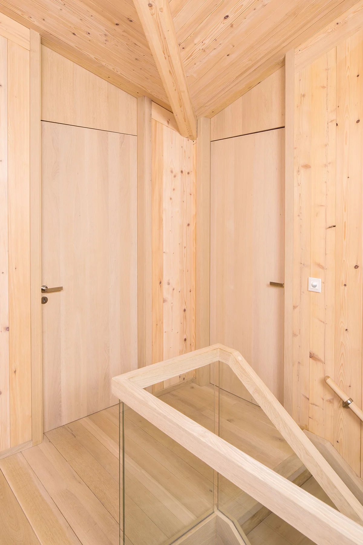 Detailaufnahme im Obergeschoss mit hellen Holztüren, hellen Massivholzwänden und sichtbarem Dachstreben. Modernes Glas-Holzgeländer und Holzfußboden. © Brunthaler Naturhausmanufaktur
