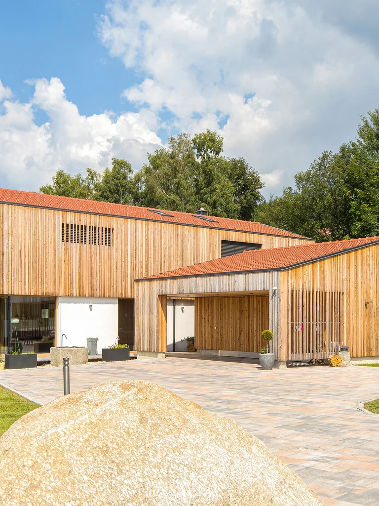 Modernes Architektenholzhaus mit vertikaler heller Holzfassade und verputztem Erdgeschoss. Ein großer überhängender Balkon und eine große Garage mit gleicher Fassade sind sichtbar. Das Haus liegt einzeln umgeben von Bäumen, Rasen und einer Einfahrt. © Brunthaler Naturhausmanufaktur