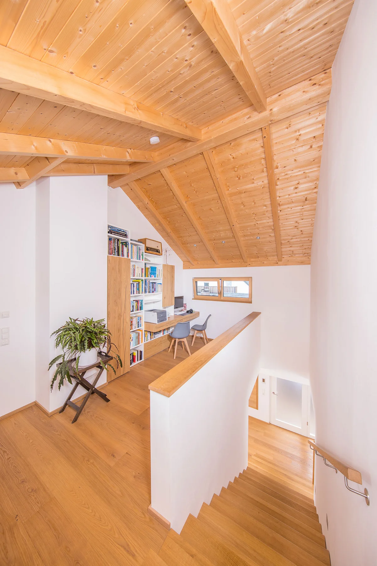Obergeschoss eines modernen Holzhauses mit Sicht auf den Dachstuhl, verputzte Wände und eine Holztreppe. Eine Ecknische ist als Homeoffice eingerichtet. © Brunthaler Naturhausmanufaktur