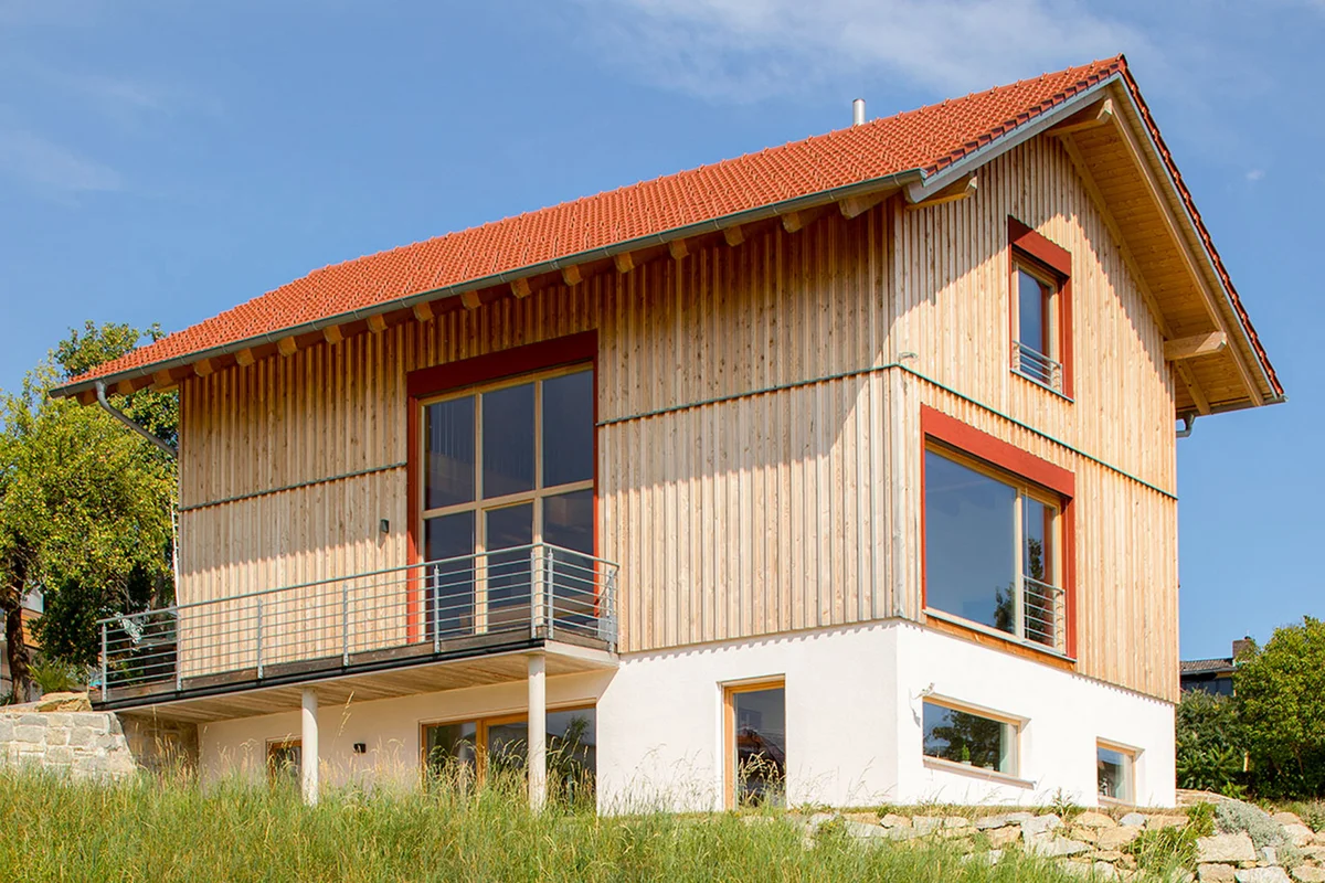 Modernes Holzhaus in Hanglage mit vertikaler Holzfassade, roten Farbakzenten, einer Holzterrasse, die sich seitlich um das Haus zieht. Der untere Bereich der Fassade ist weiß verputzt. Seitlich hat das Haus ein mehrstöckiges Fenster. Das Haus hat ein rotes Satteldach. © Brunthaler Naturhausmanufaktur