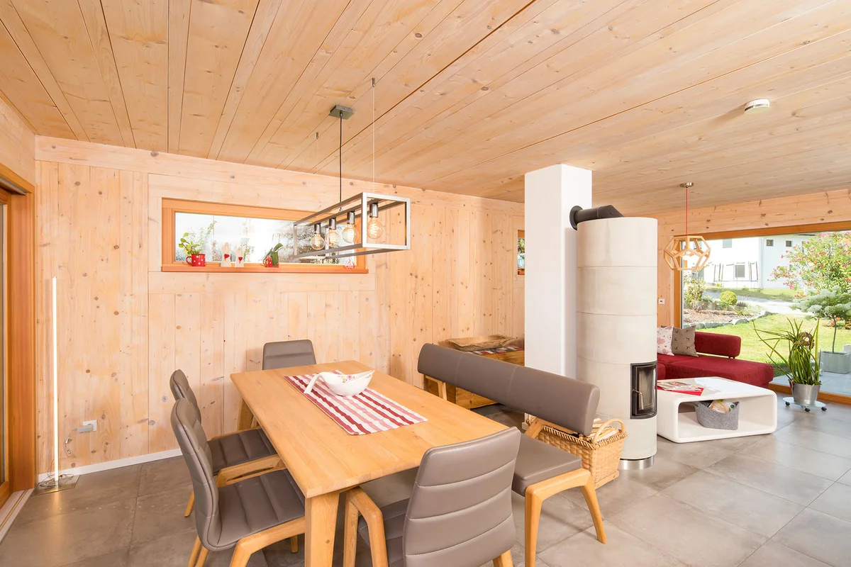 Wohn- und Essbereich in einem Holzhaus mit Holzwänden und -decken, großem bodentiefen Fenster zum Garten und einem Holzofen mit Betonoptik. © Brunthaler Naturhausmanufaktur