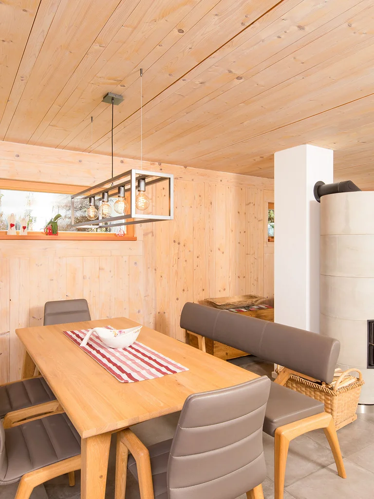 Wohn- und Essbereich in einem Holzhaus mit Holzwänden und -decken, großem bodentiefen Fenster zum Garten und einem Holzofen mit Betonoptik. © Brunthaler Naturhausmanufaktur