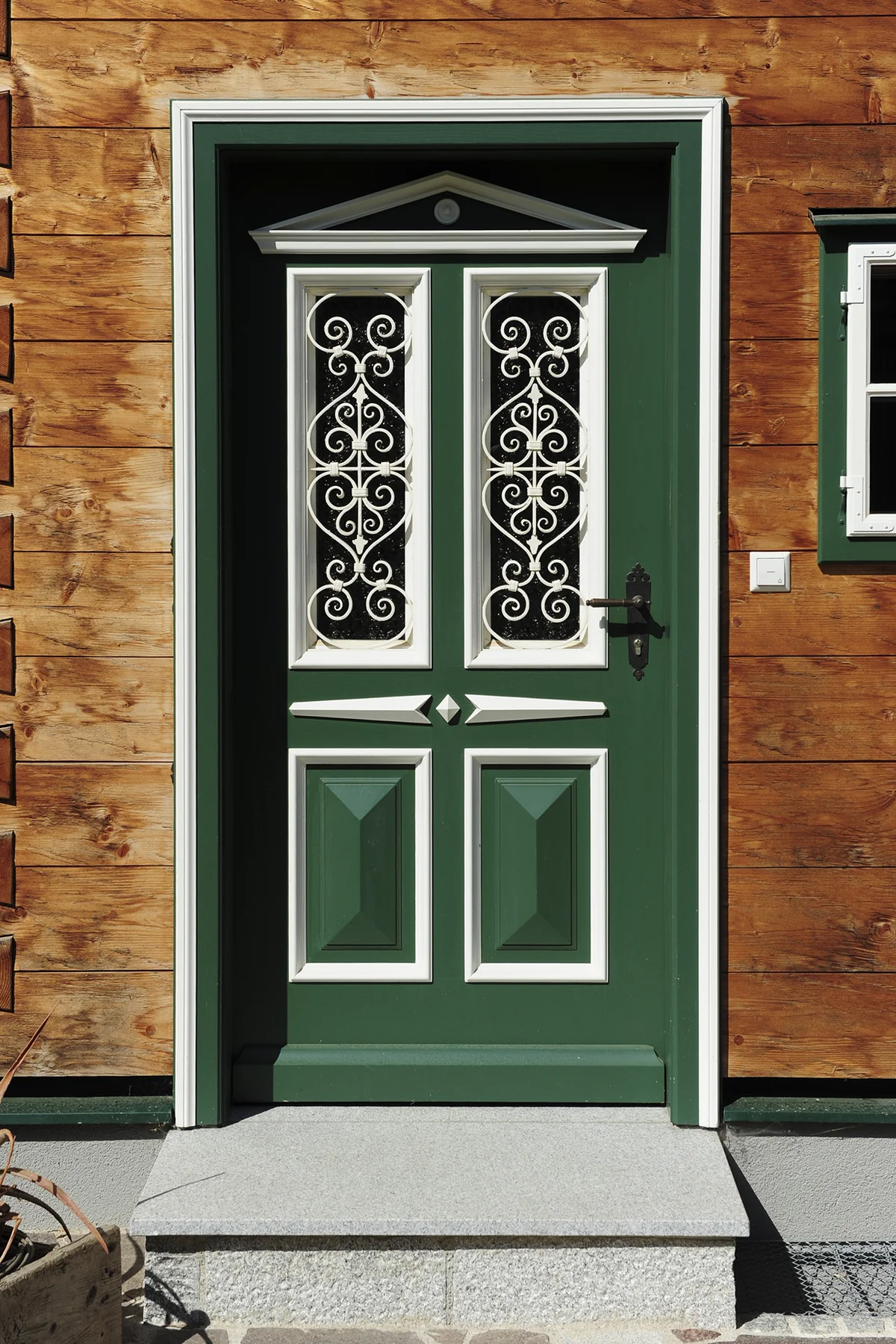 Detailaufnahme einer urig gestalteten grünen Haustür mit weißen Zierelementen und kunstvollen Gitterelementen in einer Blockbohlenfassade. © Brunthaler Naturhausmanufaktur