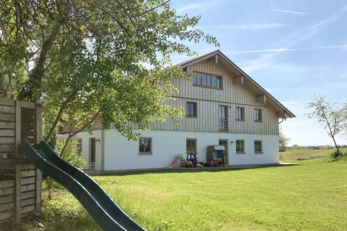 Die Rückansicht des bayerischen Landhauses mit klassischer vertikalen Holzfassade oben und verputzter Fassade unten. Es steht alleine auf einer Wiese, ein Baum und eine Kinderrutsche sind im Vordergrund. © Brunthaler Naturhausmanufaktur