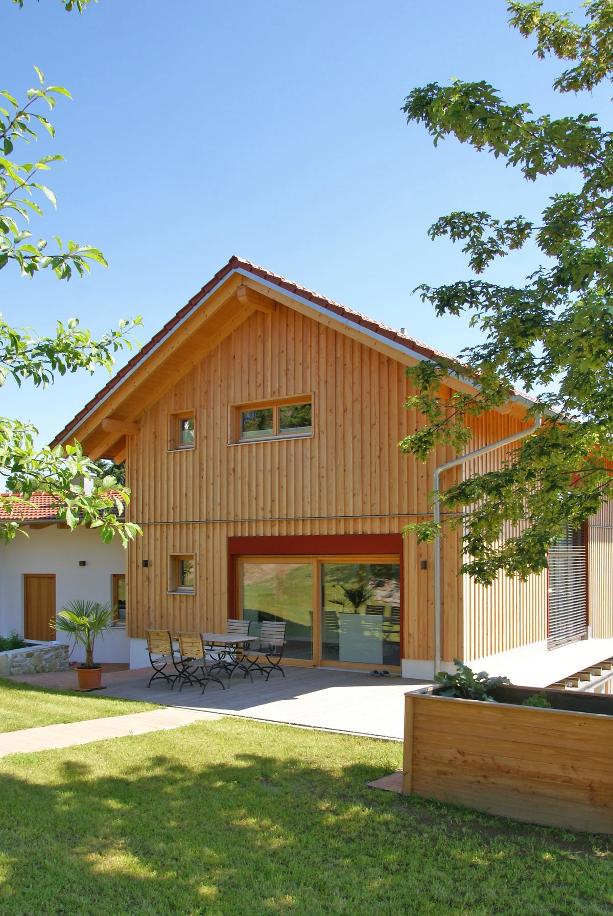 Modernes Holzhaus in Hanglage mit vertikaler Holzfassade, roten Farbakzenten, einer Holzterrasse. Es steht auf einem Grundstück mit Rasen, Bäumen und einem Hochbeet. © Brunthaler Naturhausmanufaktur