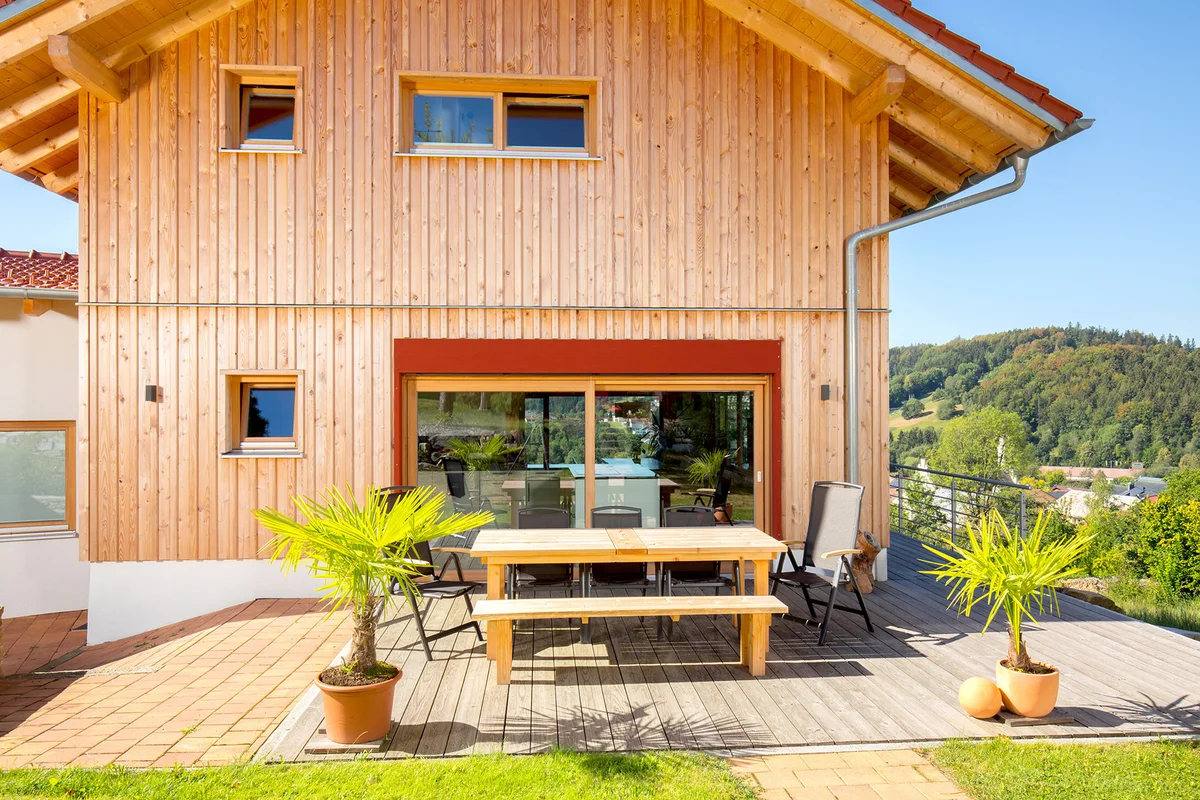Modernes Holzhaus in Hanglage mit vertikaler Holzfassade, roten Farbakzenten, einer Holzterrasse. © Brunthaler Naturhausmanufaktur