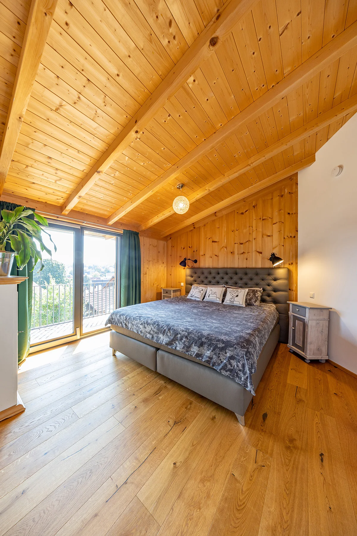 Schlafzimmer im Holzhaus mit hohen Holzdecken und teilweise verputzten Wänden. Ein großes graues Doppelbett steht im Raum mit Balkontüren und Sicht auf die Umgebung. © Brunthaler Naturhausmanufaktur