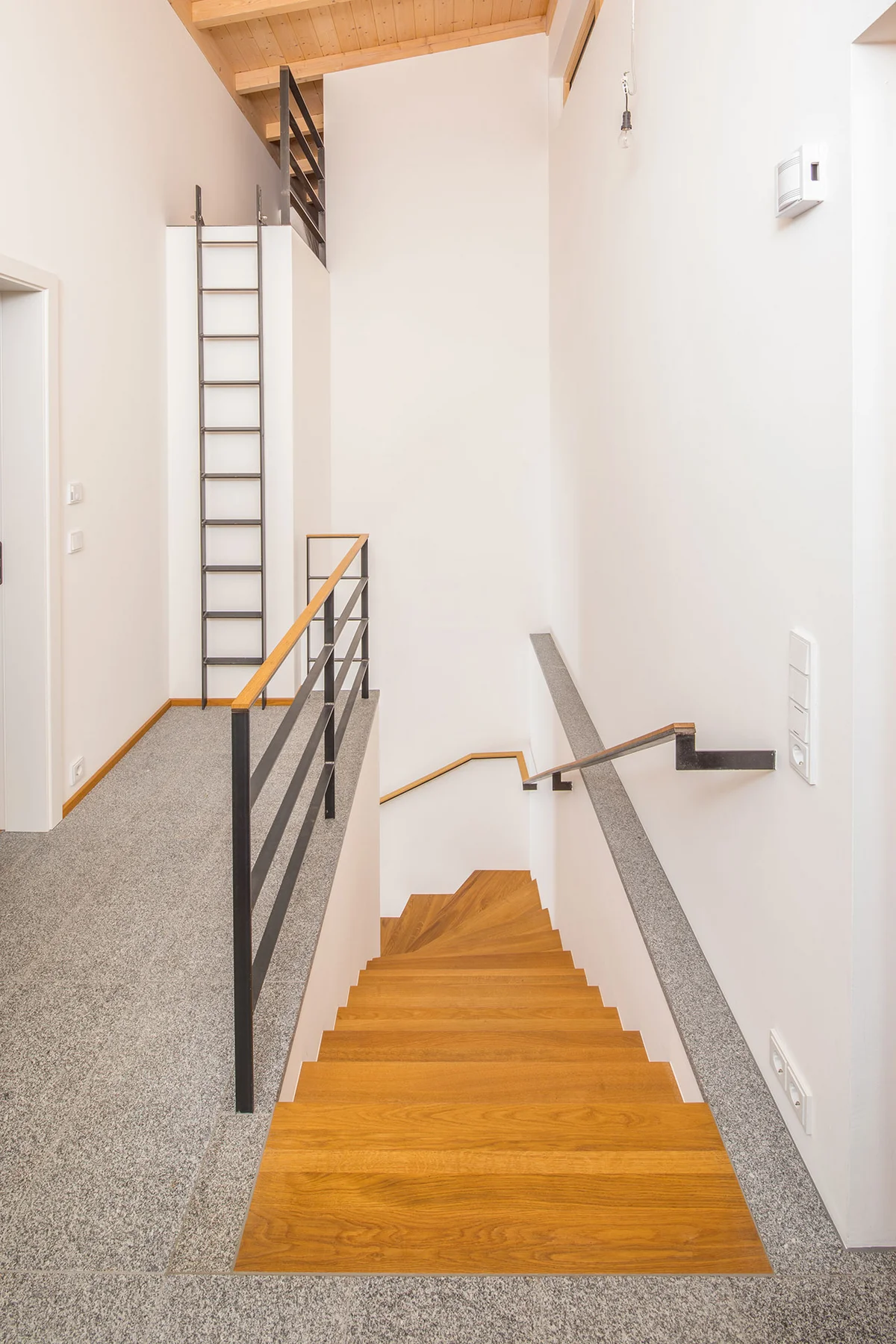 Treppenaufgang in einem Holzhaus. Eine moderne Holztreppe, drumherum weiße Wände, der Boden ist aus grauem Granit. Man sieht im hinteren Bereich eine Leiter, die über einen Vorsprung den Zugang zum Dachboden ermöglicht. © Brunthaler Naturhausmanufaktur