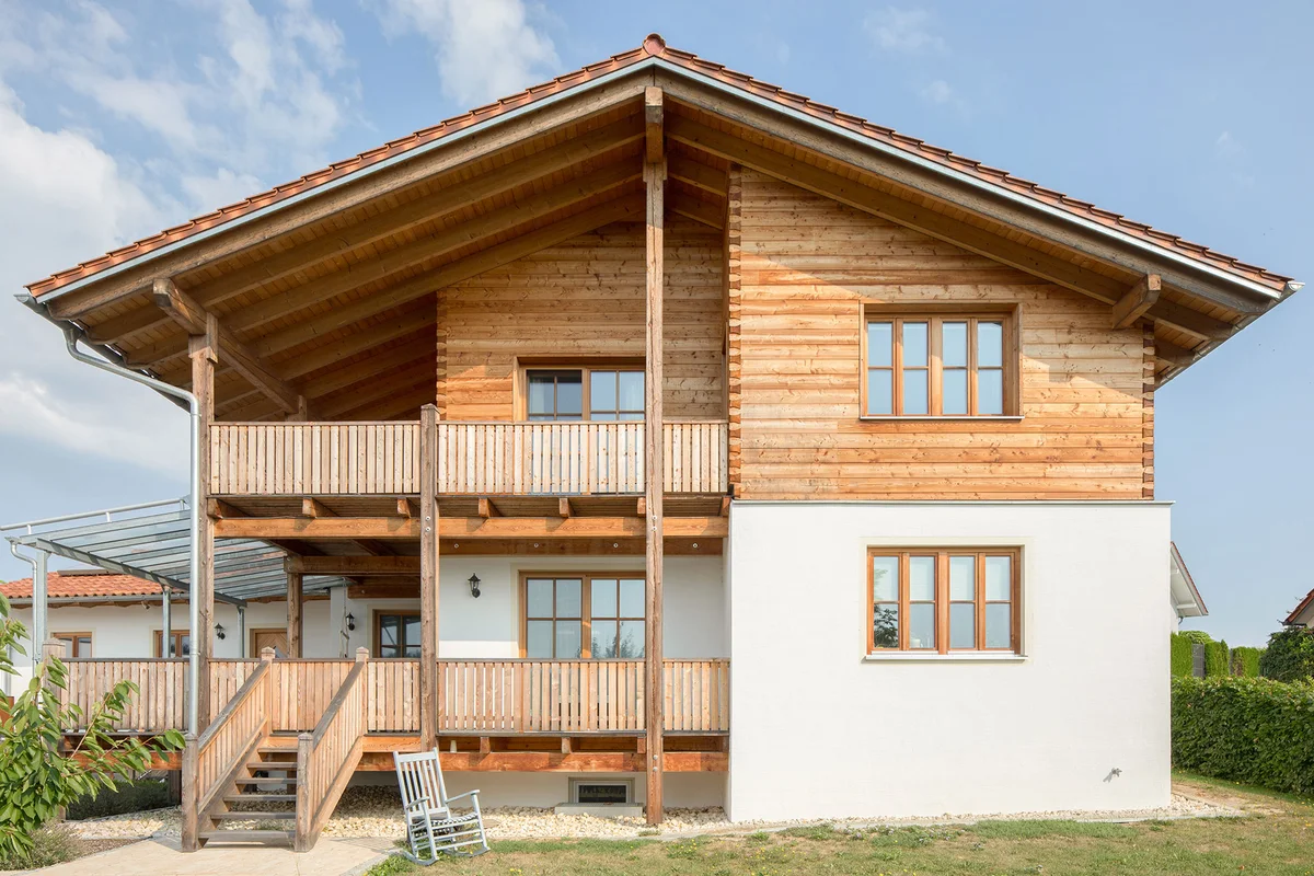 Frontale Sicht auf das Massivholz-Landhaus mit geteilter Blockbohlenfassade oben und verputzter Fassade unten. Fenster mit Holzsprossen aus naturbelassenem Holz, ebenso die Holzterrasse und der Holzbalkon. Ein hellblauer Schaukelstuhl steht im Rasen. © Brunthaler Naturhausmanufaktur
