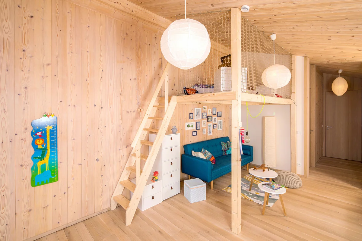 Kinderzimmer im Massivholzhaus mit Holzwänden und eine Zwischeneben als eine Art geräumiges Hochbett mit Netz und Holztreppe. Unten ist eine blaue Couch und ein Tisch und oben ist das Bett. © Brunthaler Naturhausmanufaktur