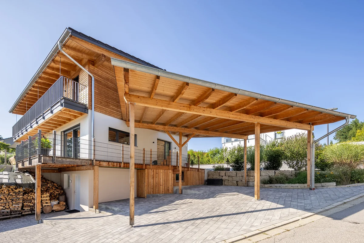 Ein Holzhaus in Hanglage mit umlaufendem Balkon, schräge gepflasterte Zufahrt mit großer Überdachung. © Brunthaler Naturhausmanufaktur