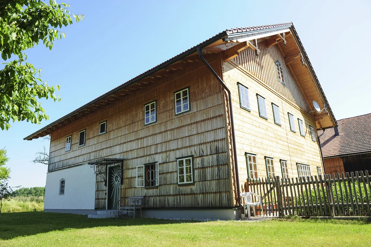 Innviertler Hof aus Massivholz mit traditioneller Holzschindelfassade, weiß verputztem Erdgeschoss und rotem Satteldach inmitten einer grünen Wiesenlandschaft. © Brunthaler Naturhausmanufaktur