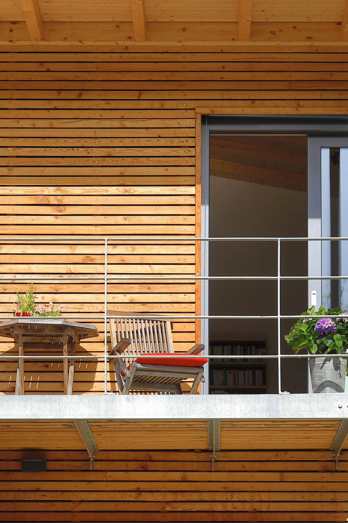 Detailaufnahme des Balkons eines Holzhauses mit unbehandelter Holzfassade und offener Balkontür sowie ein paar Holzstühlen mit roten Sitzkissen und ein kleiner Tisch. © Brunthaler Naturhausmanufaktur