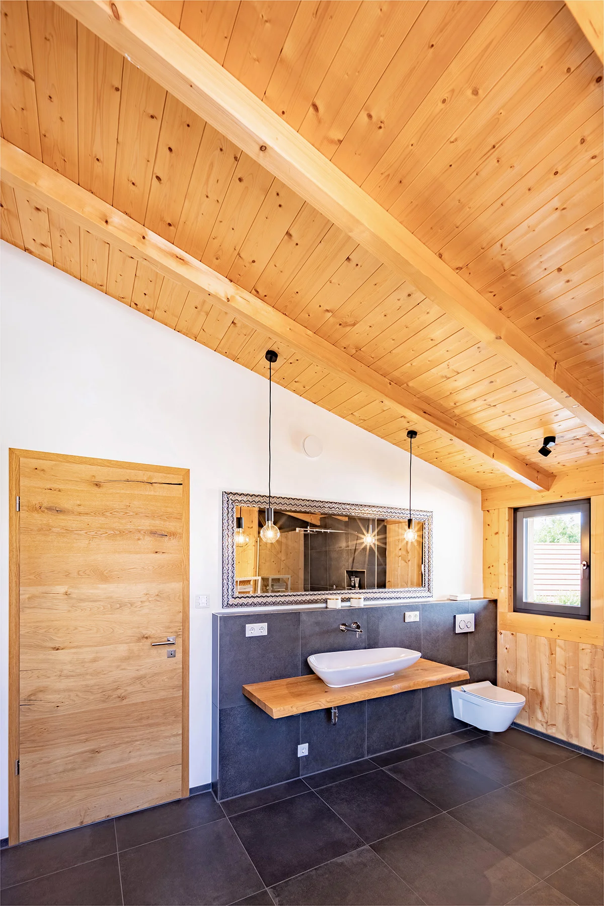 Ein Badezimmer im Holzhaus mit verputzter Wand und Holzdecke. Dunkelgraue große Fliesen sind am Boden und an der Vorwand hinter dem Klo und dem Waschbecken, das auf einem fliegenden Holztisch sitzt. © Brunthaler Naturhausmanufaktur